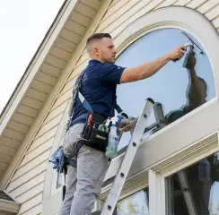 Professional window cleaner on a ladder using a squeegee to clean an exterior arched window, Santa Ana, CA.